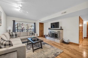 Una sala de estar con sofá, mesa de centro, chimenea, televisor y ventanas amplias.