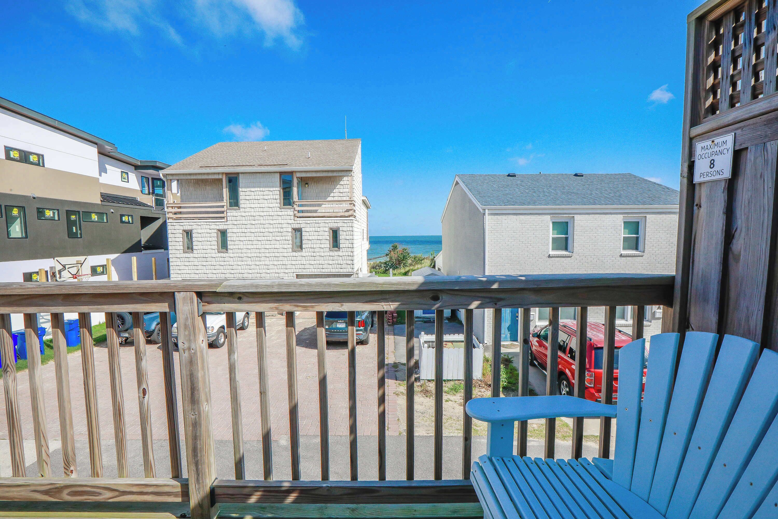 A wooden deck with a blue Adirondack chair, a view of the ocean, and a sign indicating a maximum occupancy of 8 persons.