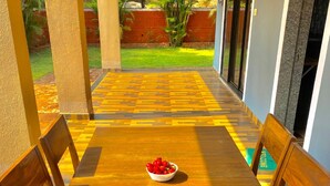 Un tavolo da pranzo in legno con una ciotola di frutta al centro, circondato da sedie, in una zona esterna coperta con pavimento decorato e vista su un giardino.