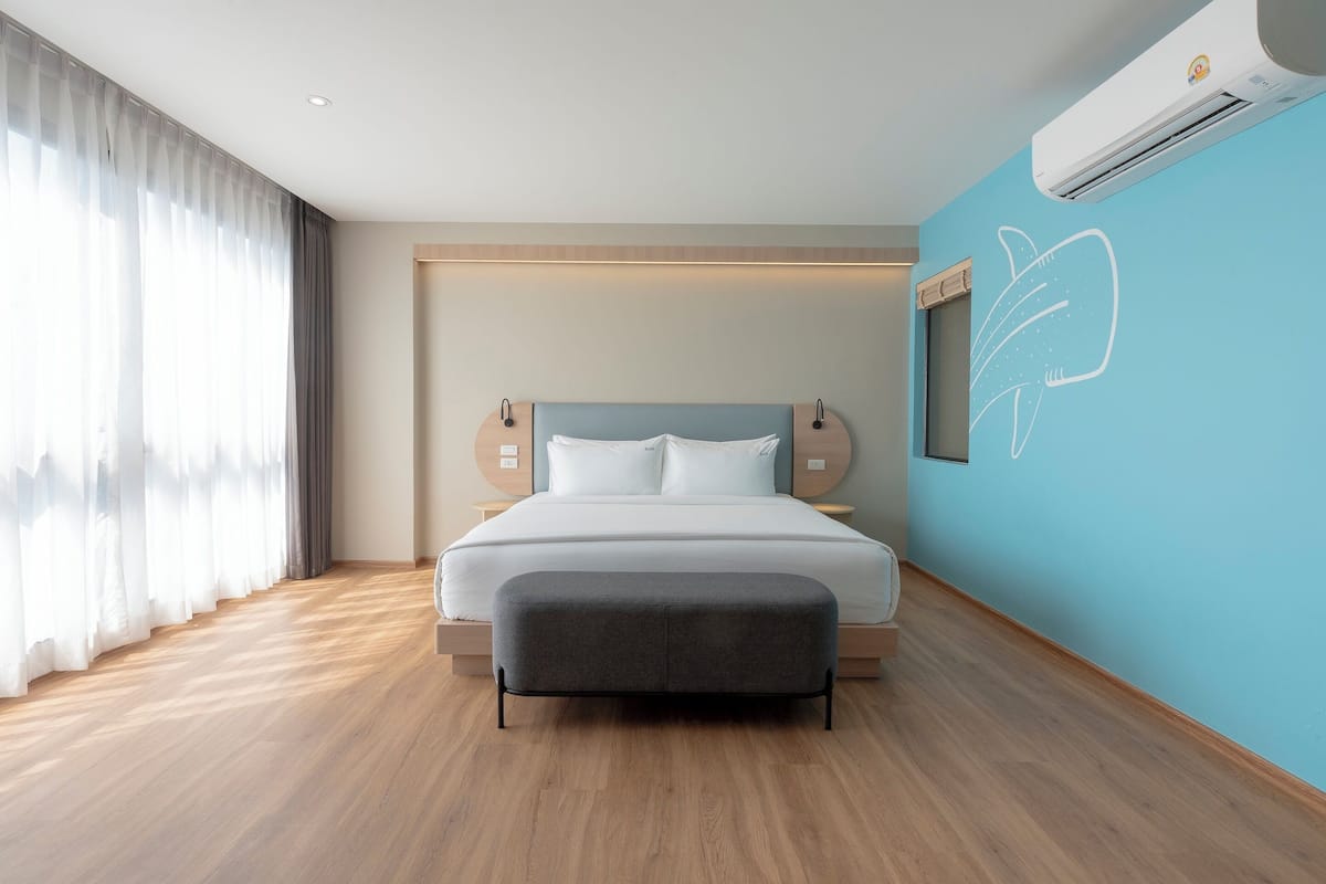 Une chambre d’hôtel moderne avec un grand lit, un pouf gris, un climatiseur fixé au mur et une fenêtre ornée de rideaux légers.
