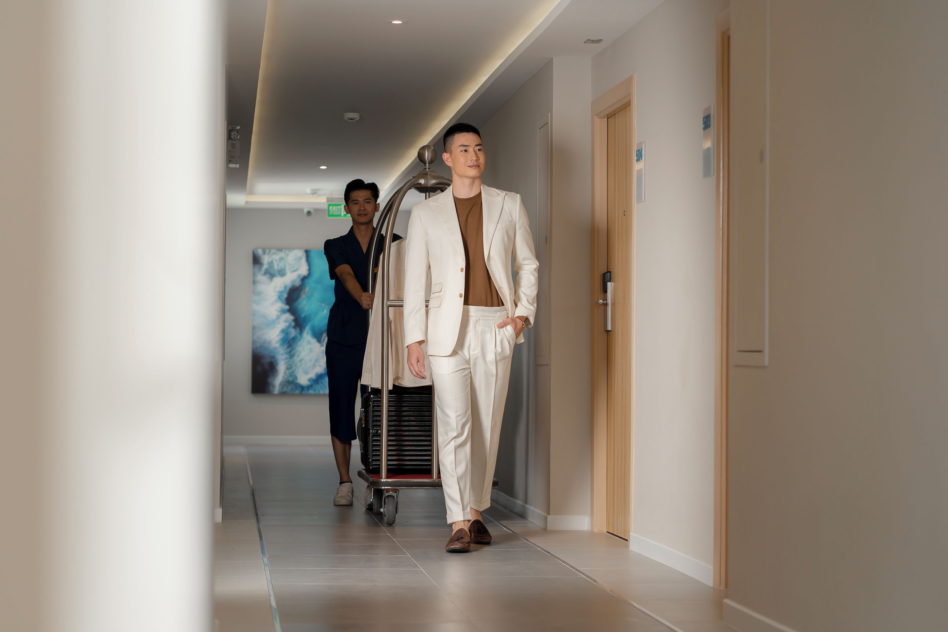 A modern hotel corridor with a person in a white suit walking, another person in a blue uniform, and a cart with luggage.
