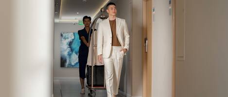 A modern hotel corridor with a person in a white suit walking, another person in a blue uniform, and a cart with luggage.