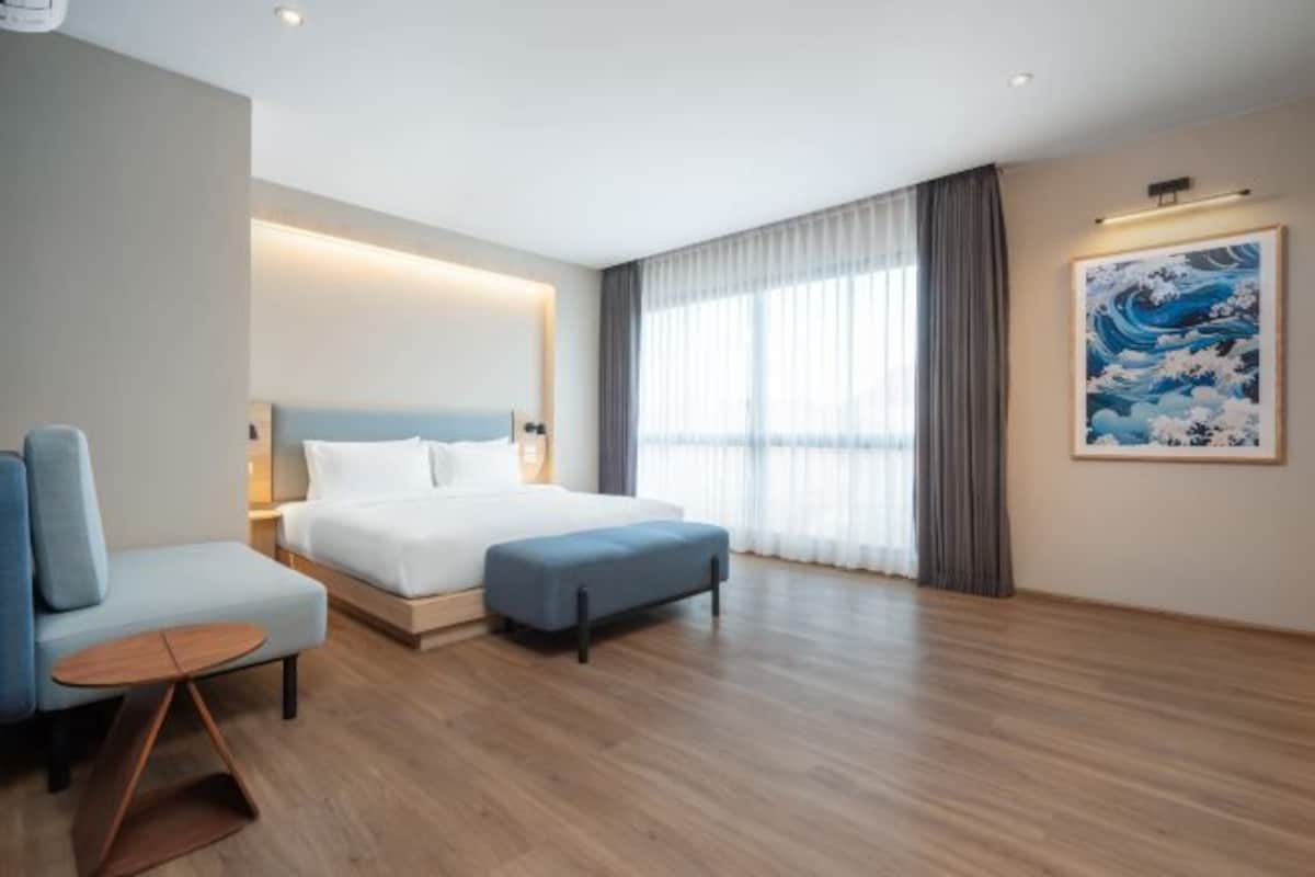 Una habitación de hotel moderna con una cama, una zona de estar, un cuadro y un ventanal con cortinas.