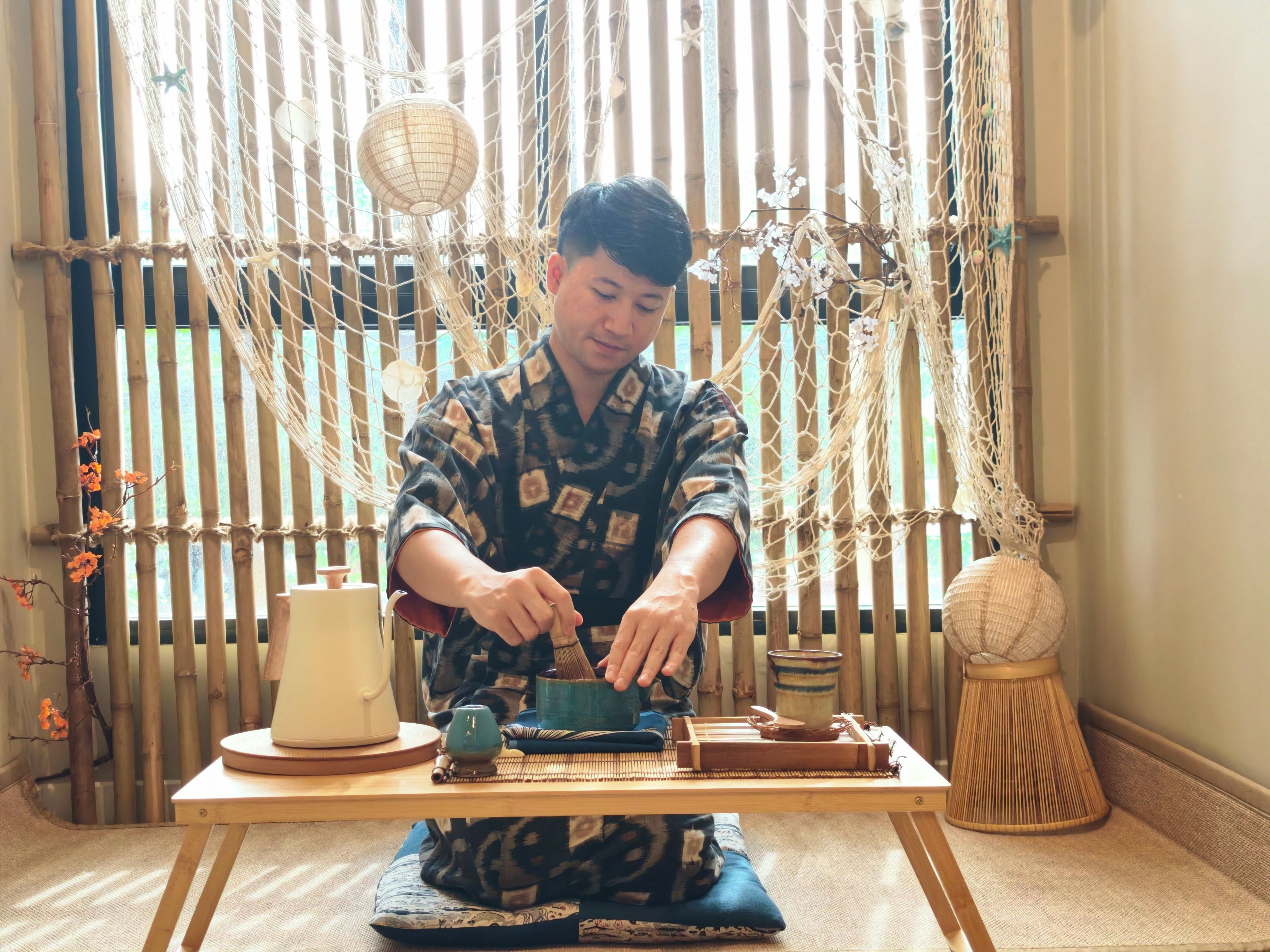 Een persoon zittend aan een tafel met een traditionele theeset, een geweven mat en een bamboebekleding.