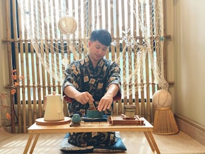 Una persona sentada a una mesa con un juego de té tradicional, un tapete tejido y un fondo de bambú.