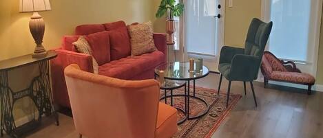 Uma sala de estar com um sofá vermelho, uma poltrona laranja, uma poltrona verde, uma mesa de centro de vidro e uma luminária.