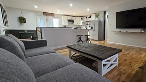 Ein modernes Wohnzimmer mit grauen Sofas, einem Holztisch und einer Küche im Hintergrund.