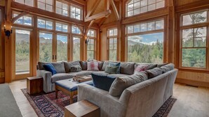 Un grand et confortable salon, doté d’un canapé d’angle, d’un plafond en bois et de grandes fenêtres offrant une vue sur les montagnes.