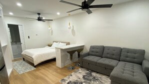 Een moderne slaapkamer met een bed, een bureau, een grijze bank, een plafondventilator en een zichtbare badkamer op de achtergrond.