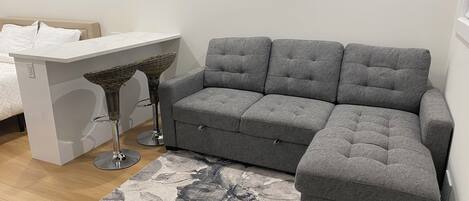 A grey sectional sofa, a white side table, and two wicker bar stools in a modern room with a patterned rug.
