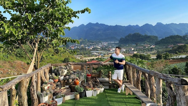A person standing on a wooden deck overlooking a lush garden with various potted plants and a view of distant mountains and a town. - Doi Thong Moc Chau - by BAY LUXURY (Moc Son)