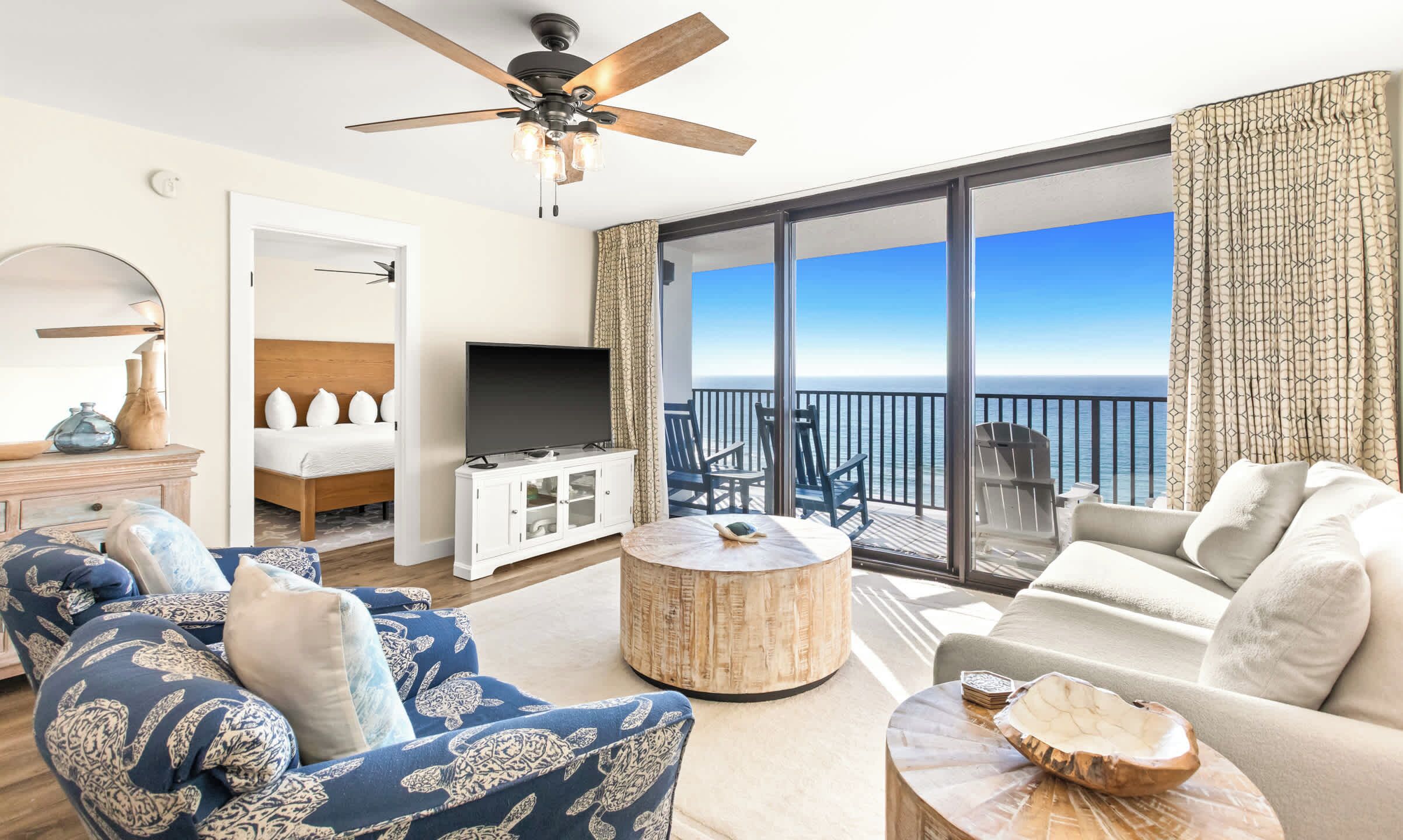 A spacious living room with a large TV, a ceiling fan, and a view of the ocean through large sliding doors.