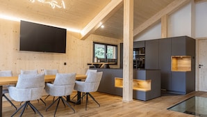 Un intérieur moderne avec un plafond en bois, un téléviseur à écran plat, un coin repas avec des chaises grises et une cuisine avec des meubles gris.