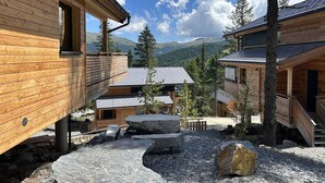 Une cabane en bois avec un sentier de pierres menant à un espace de détente, entourée d’arbres et de montagnes.