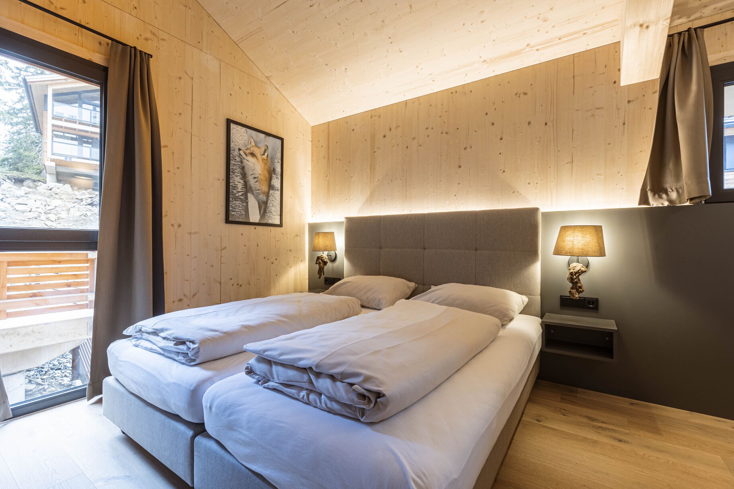 Una cama doble con cabecera, dos mesitas de noche con lámparas, un cuadro enmarcado en la pared y una ventana con cortinas.