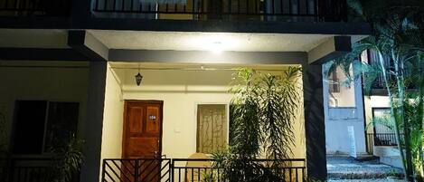A two-story building with a balcony, a door, and a small garden area with plants.