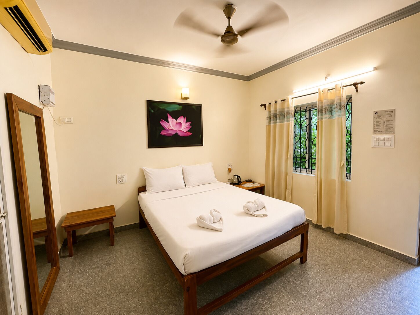 A hotel room with a bed, a bedside table, a mirror, a ceiling fan, and a window with curtains.