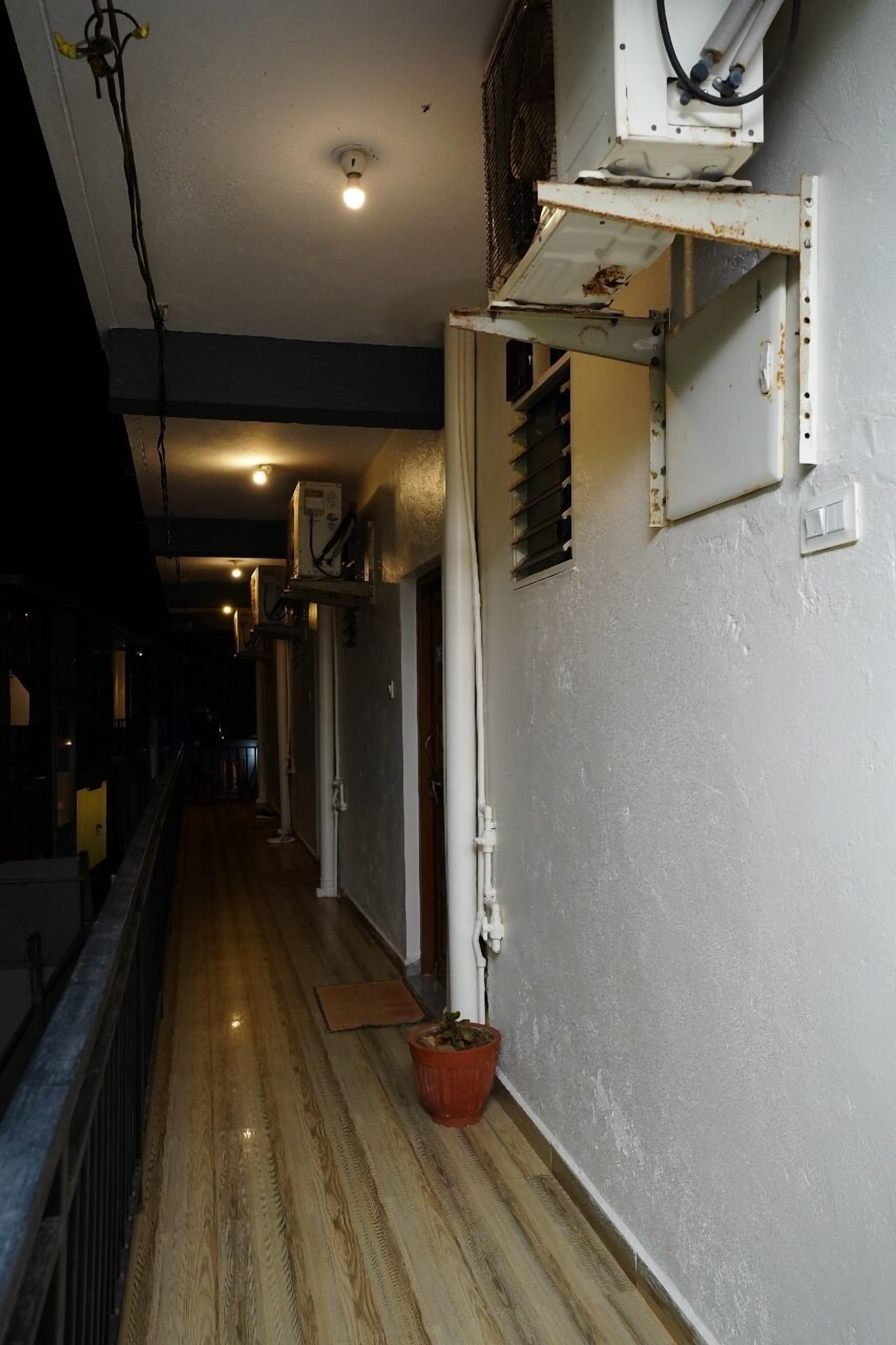 Un couloir étroit doté d’un plancher en bois, d’une plante en pot et d’un mur blanc avec une grille d’aération et un boîtier électrique.