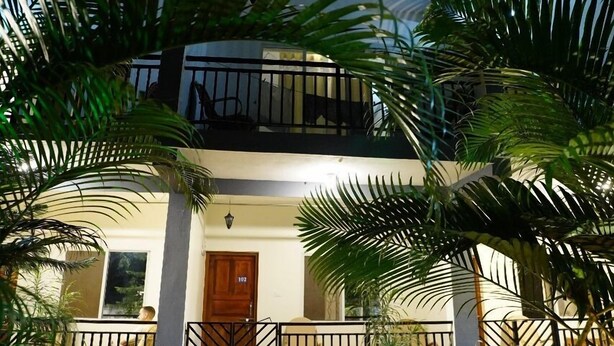 Un bâtiment à deux étages doté d’un balcon, entouré d’une végétation luxuriante et accessible par un chemin menant à l’entrée.