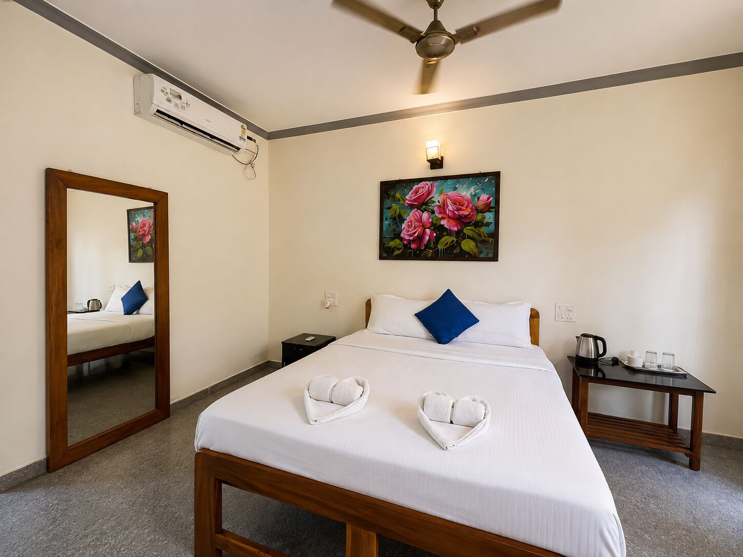 A hotel room with a bed, a nightstand, a mirror, an air conditioning unit, a ceiling fan, a painting, and a coffee table.