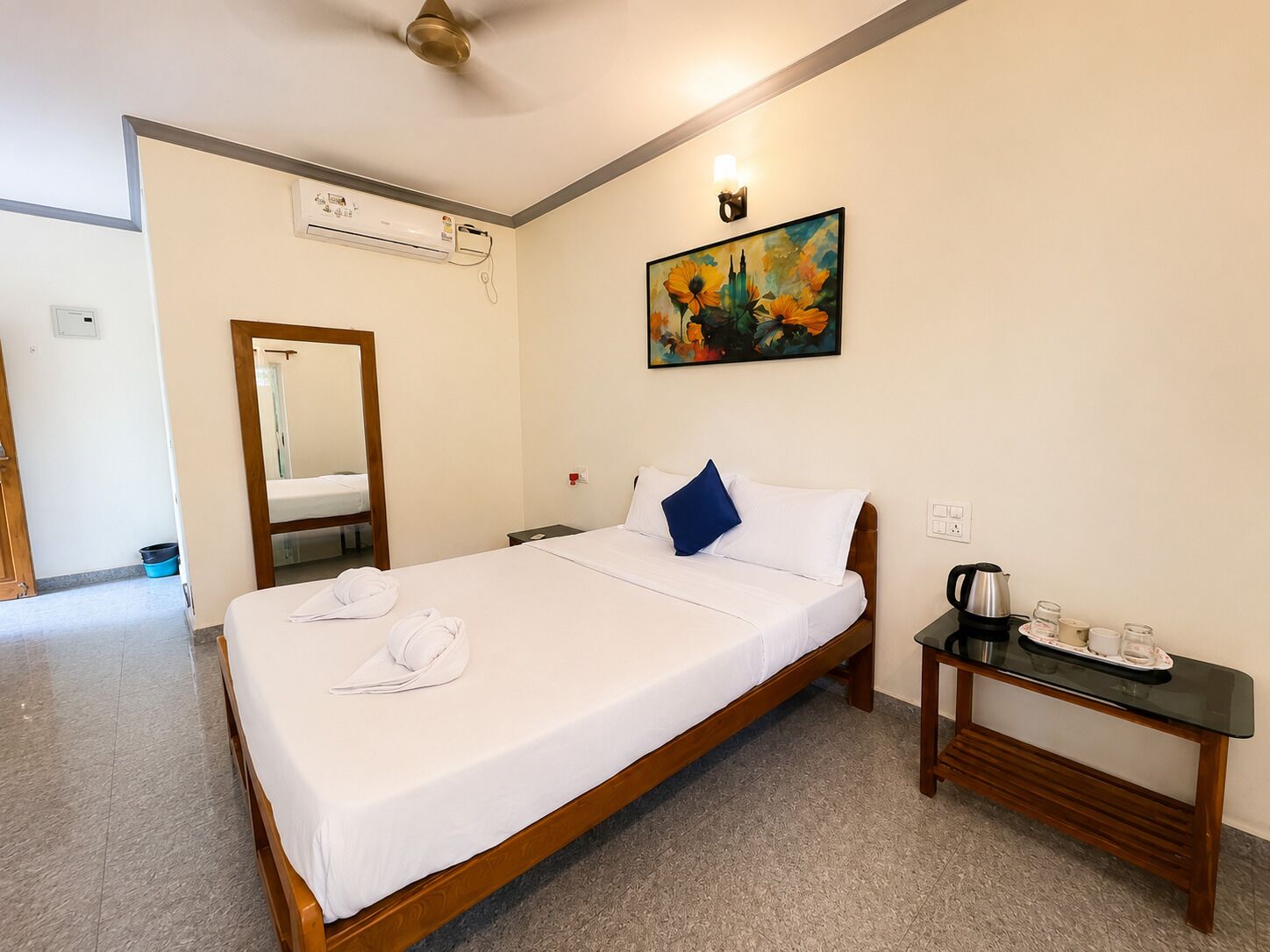 A hotel room with a bed, a bedside table with a kettle and cups, a painting on the wall, and a mirror.