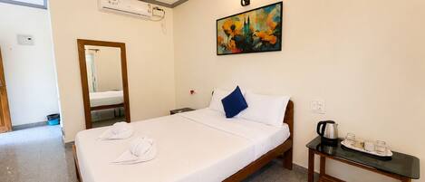 A hotel room with a bed, a bedside table with a kettle and cups, a painting on the wall, and a mirror.