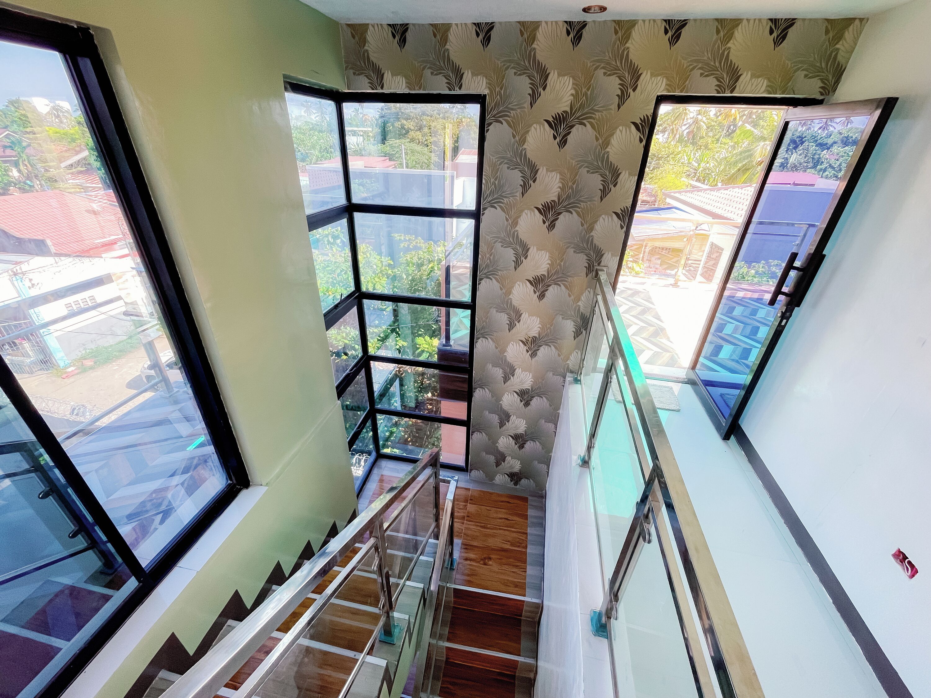 Una escalera con barandillas de vidrio, papel tapiz estampado y amplios ventanales que ofrecen vistas a edificios y zonas verdes.