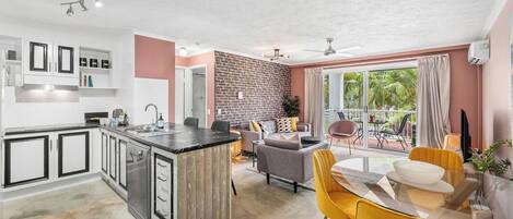 A modern kitchen with a central island, a dining area with yellow chairs, and a balcony with a view of greenery.
