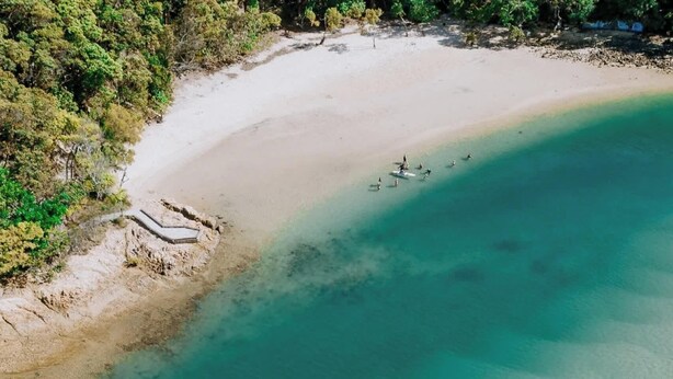 Eine Vogelperspektive zeigt einen Strand mit klarem türkisblauem Wasser und einem sandigen Ufer, auf dem sich einige Personen im Wasser befinden.