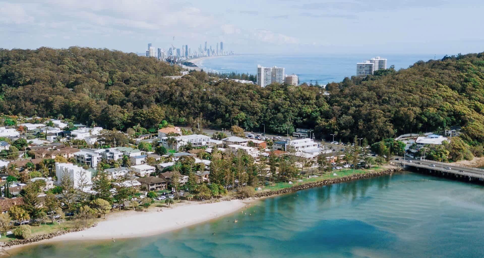 A coastal town with a beach, residential buildings, and a bridge leading to a city skyline.