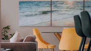 Ein Zimmer mit großem Gemälde, das Meerblick zeigt, zwei gelbe Sessel, ein graues Sofa und ein Blumentopf.