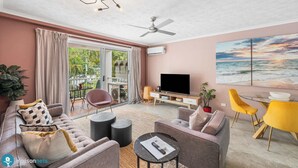 Ein modernes Wohnzimmer mit einer Couch, Sesseln, einem Couchtisch und einem Fernseher. Es gibt einen Balkon mit Blick auf Grünflächen und einen Sonnenuntergang.