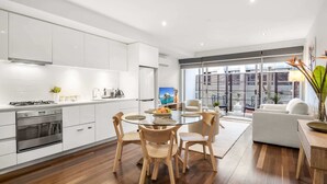 A modern kitchen with white cabinets, a dining area with a glass table and wooden chairs, and a living area with a sofa and a view of the city through large windows. - “MILA” Trendy Prahran 1-Bed Apartment with Balcony (Prahran)