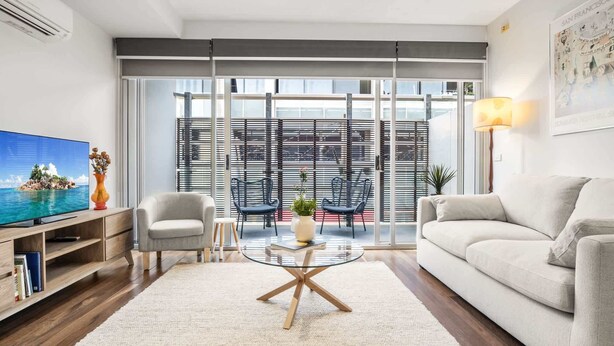 A modern living room with a sofa, armchair, and coffee table. There is a TV on a wooden stand, a lamp, and a window with blinds. - “MILA” Trendy Prahran 1-Bed Apartment with Balcony (Prahran)