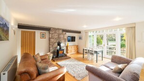 A cozy living room with a stone fireplace, a dining area with a table and chairs, and a large window offering a view of the outdoors. - The Stables Cottage Aboyne Royal Deeside (Aboyne)