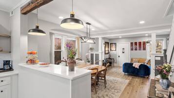 A modern kitchen with white cabinetry, a central island, pendant lighting, and a dining area with a blue sofa and wooden table.