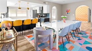 Una cocina moderna con armarios blancos, encimera negra y campana extractora de acero inoxidable. Hay una zona de comedor con una mesa blanca, sillas azules y una alfombra colorida.