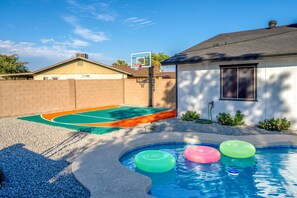 A backyard with a swimming pool, colorful inflatable rings, a basketball hoop, and a basketball court. - Backyard Game Day HQ | Pool, Hoops & Putt Putt (Glendale)
