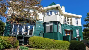 A two-story house with a green exterior and white trim, featuring a large front porch and a small front yard with trimmed bushes. - izukougen no chiisana o-beruju arusion (Ito)