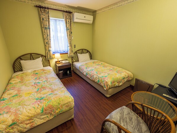 Una habitación con dos camas individuales, una mesita con lámpara, una silla y un escritorio con ordenador.