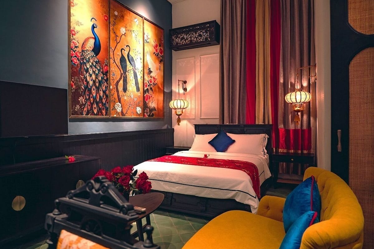 A hotel room with a bed, a nightstand, a chair, a table with flowers, and a wall with a peacock painting.