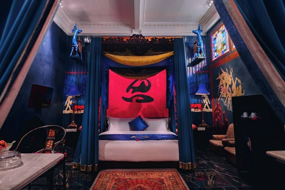 A room with a bed centered under a large red and black tapestry, blue drapes, and a chandelier.