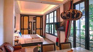 A hotel room with a bed, a TV, a dining table with a basket of fruit, and a view of greenery outside. - House of Tugu Old Town Jakarta (Jakarta)