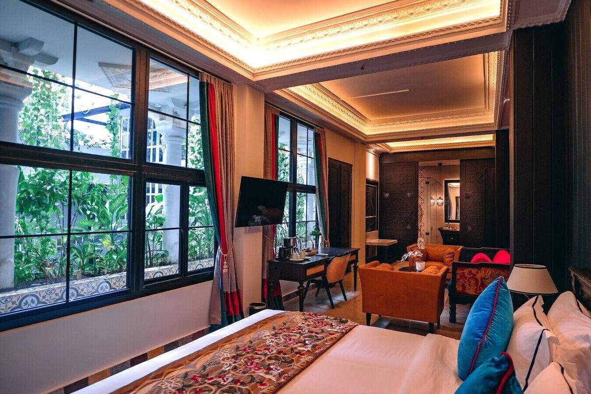 A hotel room with a large bed, a desk, a chair, and a sofa. There is a TV on the wall and a window with curtains.