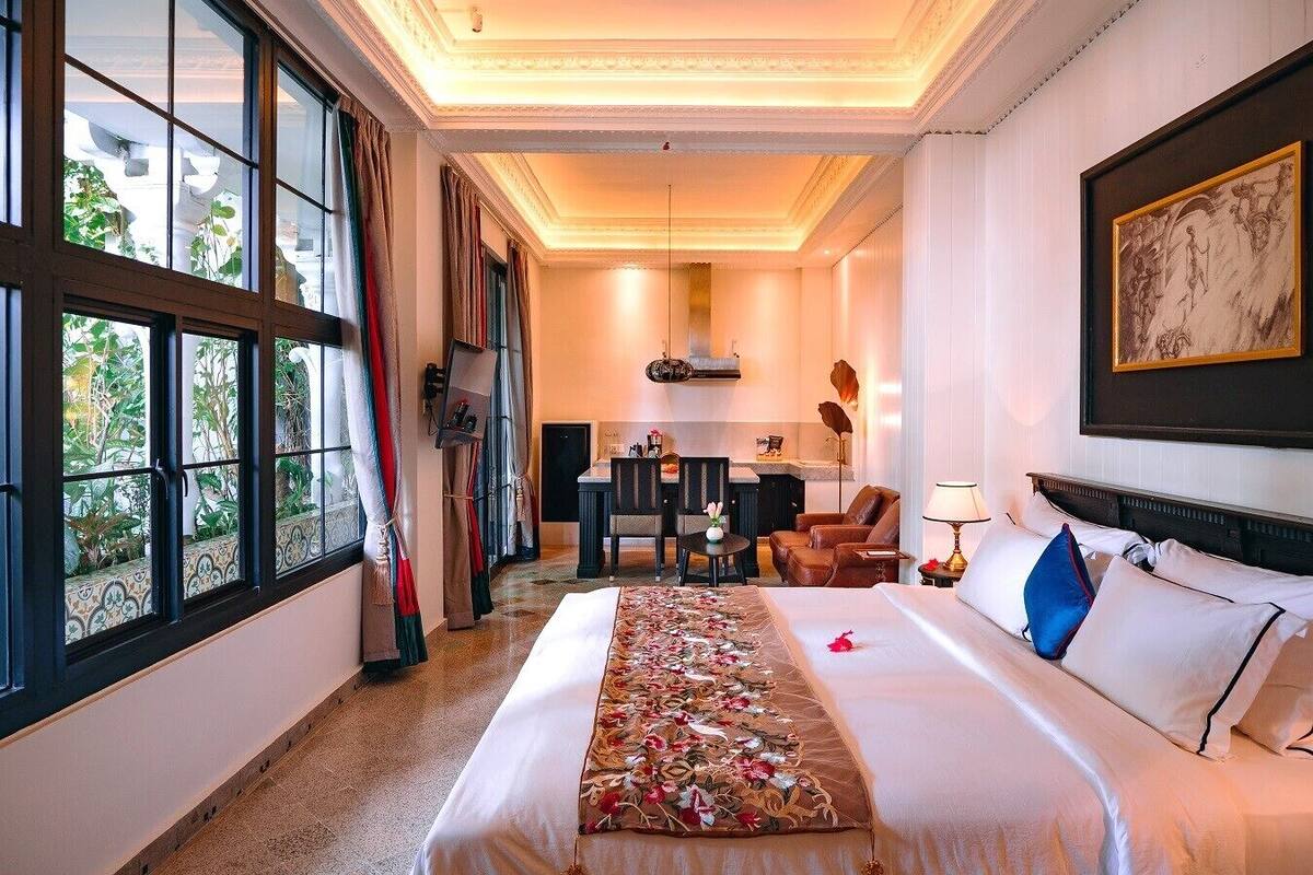 A hotel room with a large bed, a desk, a chair, and a view of greenery through the windows.
