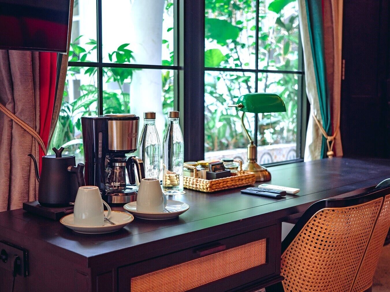 A wooden table with a coffee maker, cups, and bottles, near a window with greenery outside.