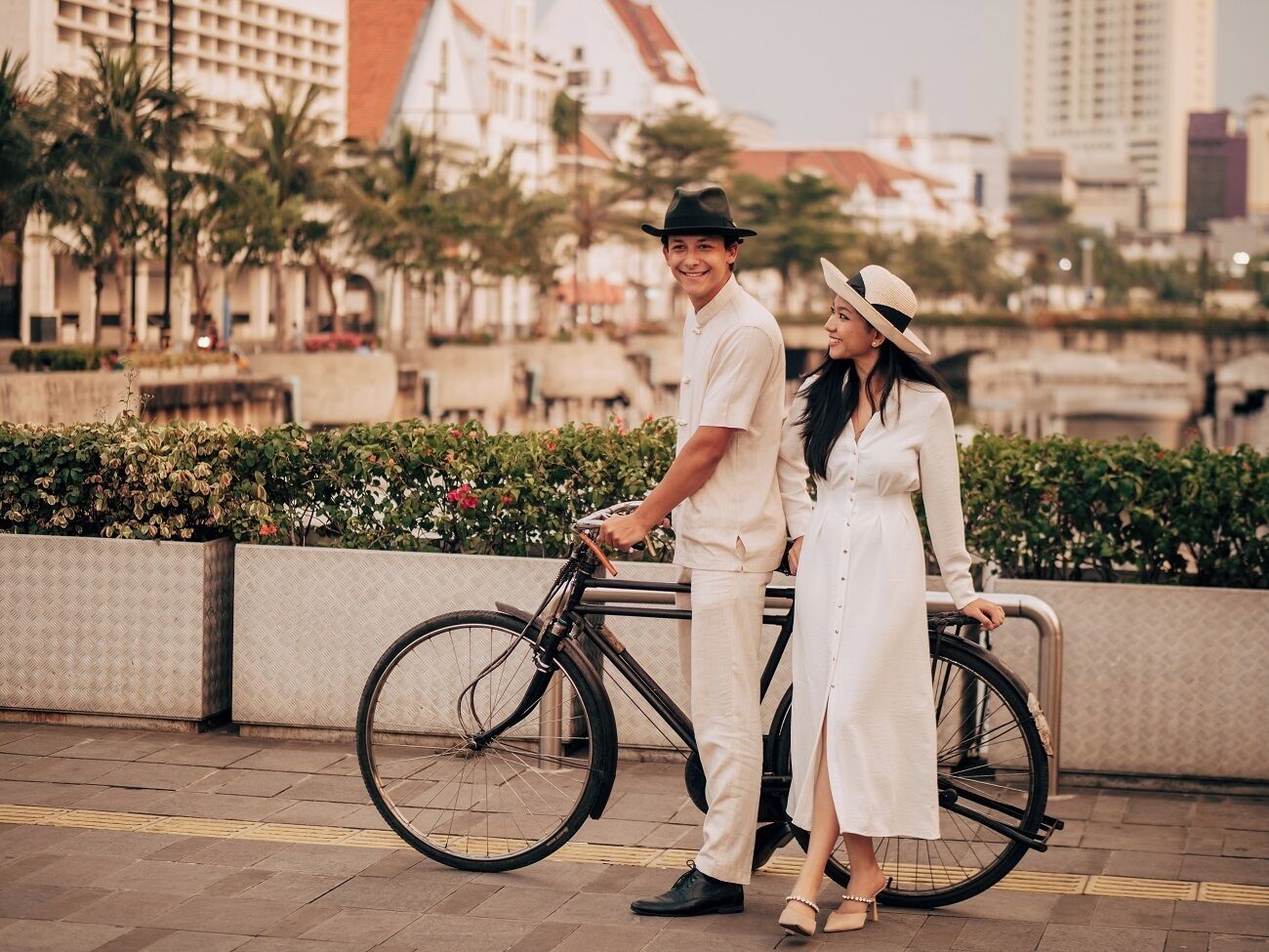A couple standing next to a bicycle in an urban setting with buildings and greenery in the background.