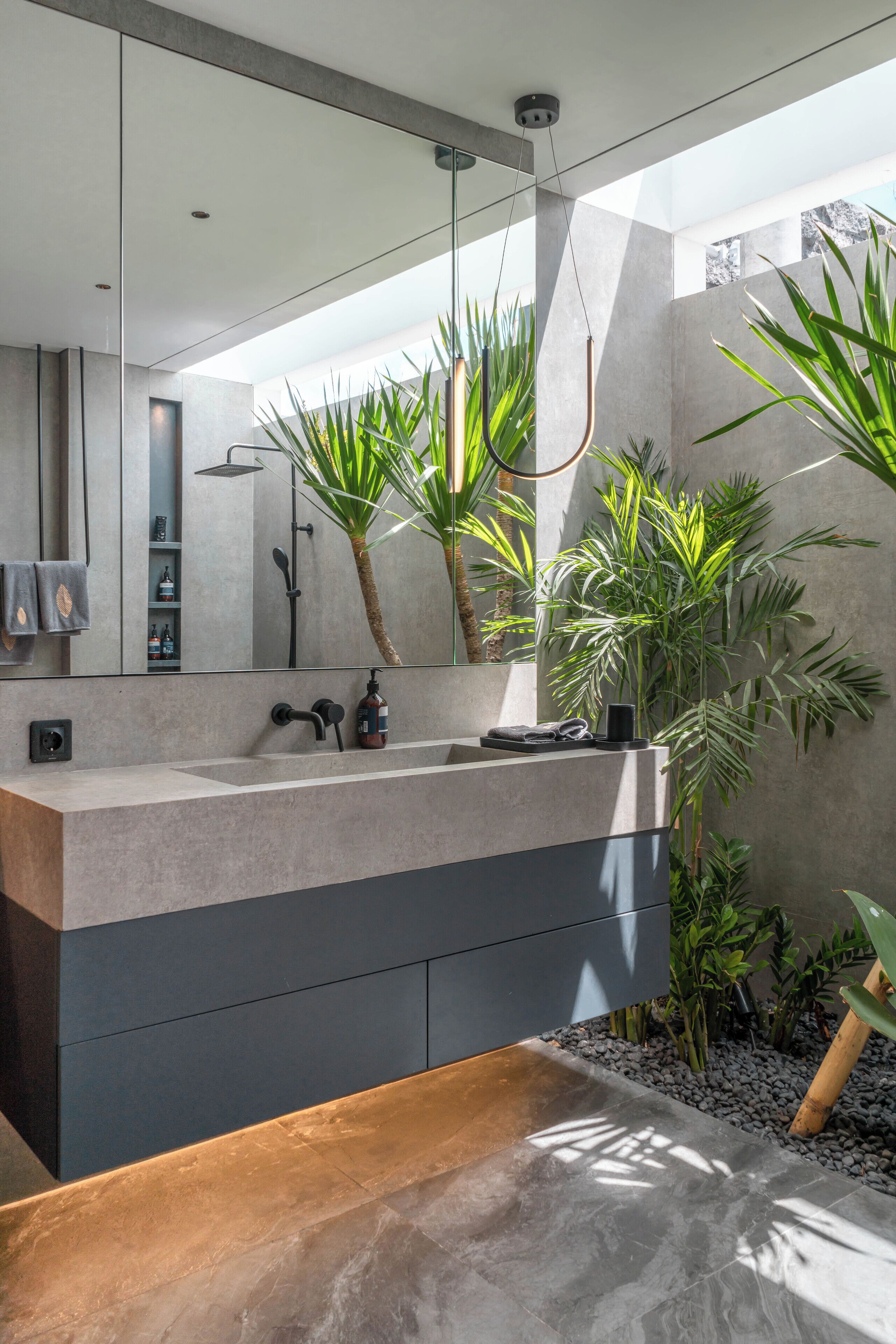 Un baño moderno con un espejo grande, un lavamanos empotrado y una ducha. Hay plantas y un banco en el espacio.