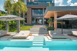 Un área de piscina moderna con sillones de descanso y sombrillas, una escalera que lleva a un edificio y abundante follaje.