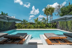 A pool area with lounge chairs and umbrellas, surrounded by lush greenery and a clear blue sky. - Beyond The Palms (Canggu)